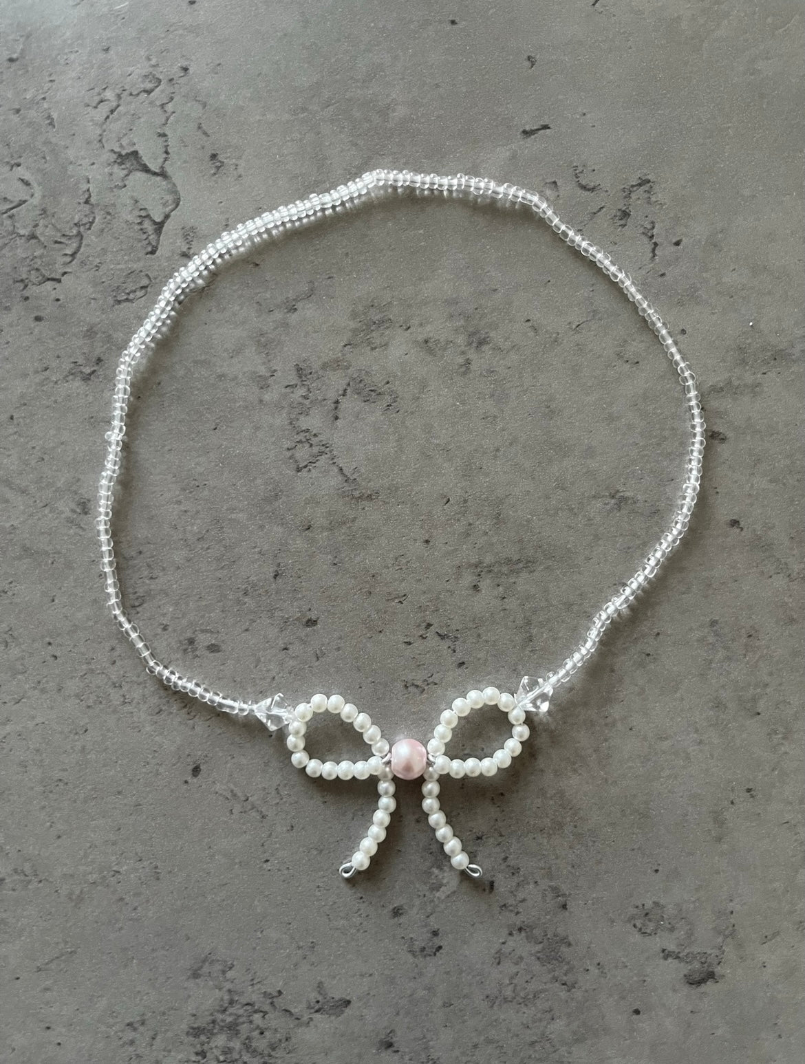 Håndlavet sløjfehalskæde med en lyserød perle i midten. Halskæden er lavet af akryl- og glasperler. Front billede.